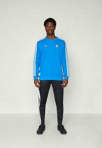 Modra trenirka z črnimi in belimi črtami, z logotipom na prsih. V kombinaciji s črnimi hlačami, ki imajo bele stranske črte.