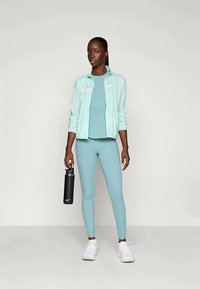 Vaaleansininen urheilutakki, jossa on vetoketjullinen etuosa, yhdistettynä yhteensopivaan yläosaan ja legginsseihin. Kädessä musta vesipullo, valkoiset lenkkarit.