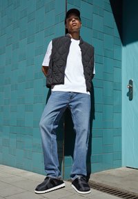 Black quilted vest over a white t-shirt, blue jeans, and black slip-on shoes with white soles, against a textured teal wall.