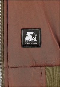 Brązowa pikowana tkanina z czarną naszywką z logo "Starter Black Label". Posiada żebrowane akcenty w oliwkowej zieleni wzdłuż krawędzi i zamka.