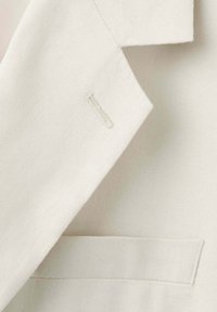 Gros plan sur le revers d’un blazer couleur crème avec une boutonnière et une poche poitrine horizontale sur tissu texturé.