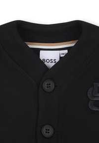 Camicia nera con colletto a V, dotata di due bottoni neri e dettagli del logo ricamato sul davanti. Tessuto morbido e liscio.