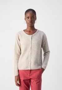 Gilet beige clair avec une texture douce, encolure ronde et six boutons sur le devant. Porté par-dessus un pantalon rose.