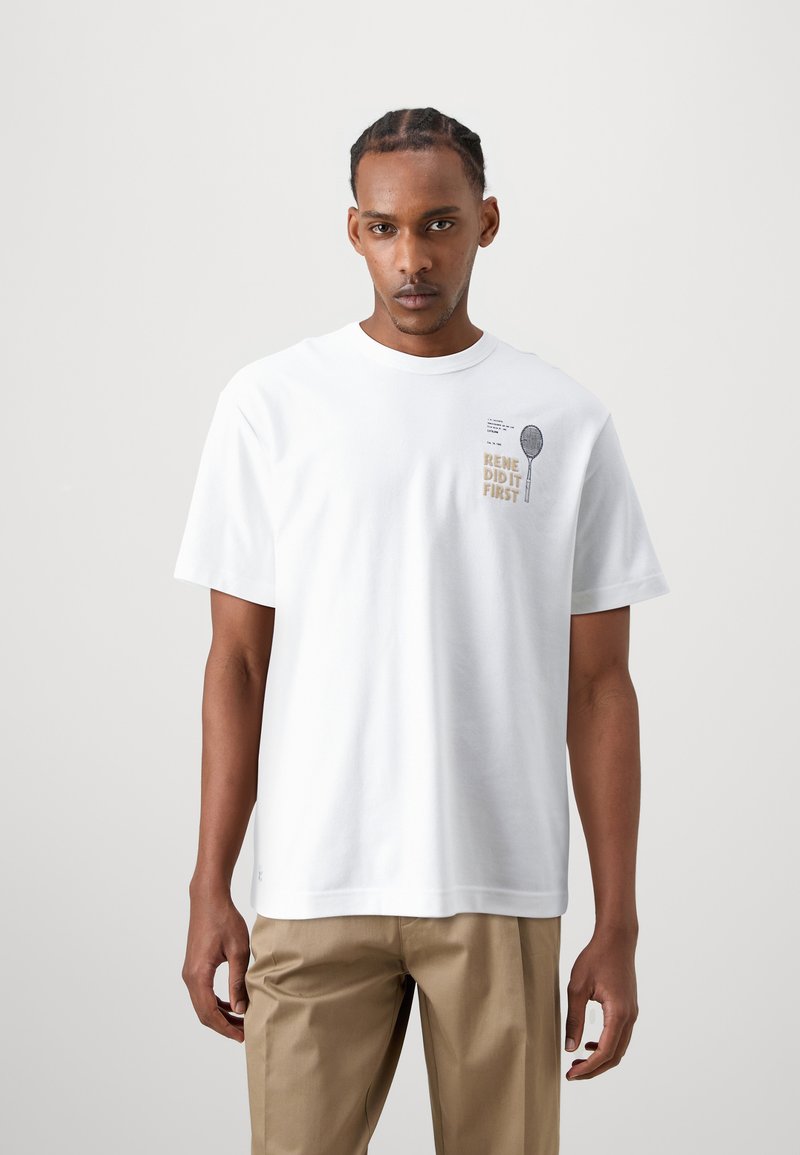 T-shirt bianca a maniche corte in cotone, con una grafica serigrafata che presenta del testo e un'immagine di una racchetta da tennis nella zona del petto sinistro.