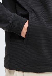 Χέρι μερικώς μέσα στην πλαϊνή τσέπη ενός μαύρου ριμπλό πλεκτού μακρυμάνικου ρούχου που φοριέται πάνω από ανοιχτό μπεζ παντελόνι.