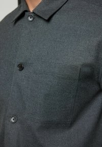 Graues, kurzärmeliges Hemd aus strukturiertem Stoff, mit einer kleinen Brusttasche und schwarzen Knöpfen. Das Design umfasst einen klassischen Kragen.