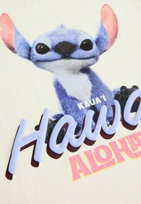 Camiseta de algodón con un personaje de dibujos animados azul con orejas grandes, texto "Hawai'i ALOHA" en rosa y azul claro, y un fondo beige claro.