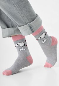 Graue Baumwollsocken mit pinken Akzenten, die ein Snoopy-Motiv und Herzmuster zeigen. Über den Socken sind aufgekrempelte Jeans sichtbar.
