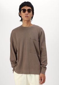 Brun langermet genser med rund hals og brystlomme, laget av mykt stoff. Modell har på seg mørke solbriller. Lys bakgrunn.