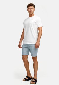 Witte katoenen t-shirt met een borstzak, gecombineerd met lichtblauwe shorts. Model draagt zwarte sandaal met verstelbare banden. Eenvoudig ontwerp.