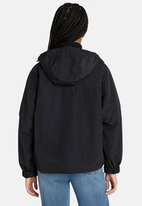 Giacca nera impermeabile con cappuccio, polsini elasticizzati e vestibilità rilassata. Presenta un logo discreto sulla parte posteriore e una texture liscia e opaca.