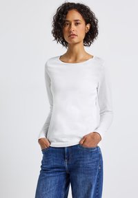 Frau mit lockigem, dunklem Haar, die ein weißes Langarmshirt und blaue Jeans trägt, steht mit den Händen in den Taschen vor einem schlichten Hintergrund.
