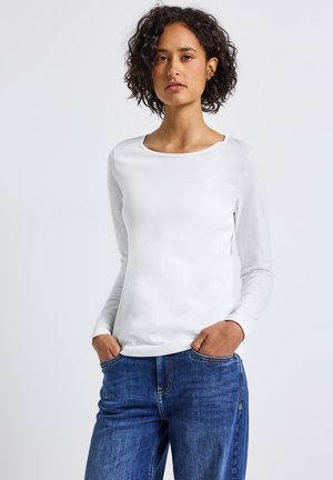 Vrouw met krullend donker haar in een witte blouse met lange mouwen en blauwe spijkerbroek, staand met handen in de zakken tegen een effen achtergrond.
