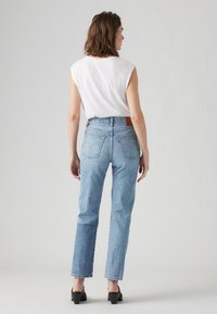 Jeans azuis de cintura alta com perna reta, bainha desfiada e bolsos traseiros. Combinados com uma blusa branca sem mangas. Sapatos de salto pretos completam o visual.