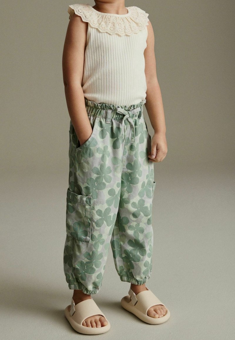 Enfant portant un haut sans manches crème à côtes avec col en dentelle, pantalon vert à imprimé floral avec poches, et sandales crème à bout ouvert, debout devant un fond uni.