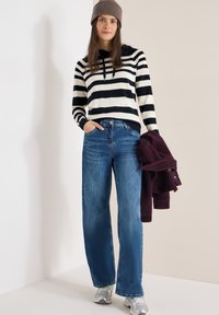 Femme portant un sweat à capuche rayé noir et blanc, un jean bleu, des baskets argentées et un bonnet en tricot marron, tenant une veste pliée de couleur violet foncé.