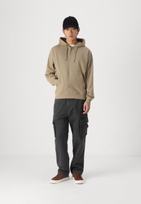 Beige bomullshuvtröja med en framficka, tillsammans med mörka cargobyxor som har flera fickor. Modellen bär bruna sneakers och en svart keps.