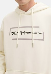 Crème-kleurige hoodie met een voorkantgrafiek die "DENIM TOM TAILOR" in zwart en grijs weergeeft. Voorzien van een trekkoordkap en een kangoeroezak.