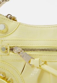 Gros plan d'un sac à main texturé jaune pâle avec des clous dorés et une fermeture éclair avec une tirette métallique et un pompon en cuir attaché.