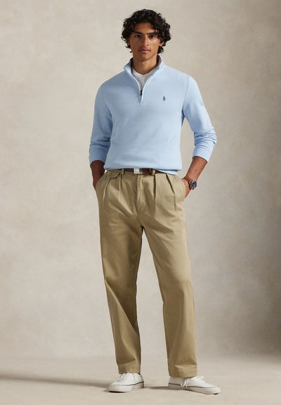 Νεαρός άνδρας όρθιος με τα χέρια στις τσέπες, φορώντας ανοιχτό γαλάζιο πουλόβερ με φερμουάρ έως τον λαιμό, μπεζ πλισέ παντελόνι, λευκά αθλητικά παπούτσια και ρολόι χειρός.