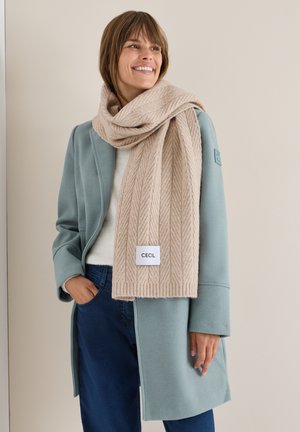 Beige, mønstret tørklæde med et fiskebensmønster og et mærke, viklet om en person iført en lyseblå frakke og en hvid sweater.
