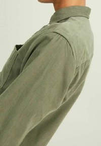 Camicia in corduroy verde oliva con colletto alto, maniche lunghe e un'unica tasca sul petto. Il tessuto testurizzato presenta righe verticali.