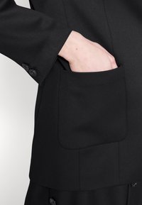 Černý blejzer s elegantním designem; má viditelnou kapsu, hladkou texturu a dva knoflíky na manžetě pro zapínání.