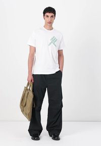 Hvid bomulds t-shirt med grøn tekst, sorte cargo bukser og en beige tote bag. Modellen står foran en neutral hvid baggrund.