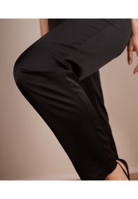 Svarta, slim-fit-byxor med en mjuk textur och skräddarsydd design. Innehåller en subtil sömdetalj längs benet, i kombination med en svart skosnodd.