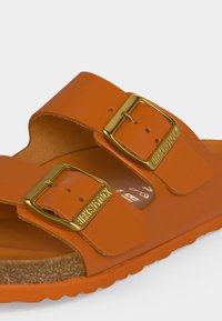 Πορτοκαλί σανδάλι Birkenstock με δύο χρυσά μεταλλικά κουμπώματα και φελό πάτο, κοντινό πλάνο που δείχνει υφή και σήμανση στα κουμπώματα.