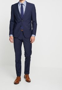 Marineblauw op maat gemaakt pak met een slim fit, voorzien van een blazer met twee knopen, notch revers en rechte broek. Gecombineerd met een lichtblauwe dress shirt en bruine leren schoenen.