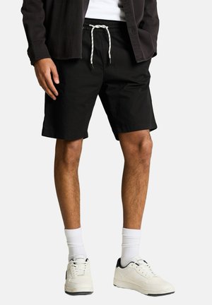 Mann trägt schwarze Shorts mit Kordelzug, weiße Socken und weiße Sneakers, steht mit einer Hand in der Tasche und der anderen an der Seite.