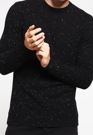 Man wearing a black speckled knit sweater, adjusting the wrist area with both hands visible against a plain background.