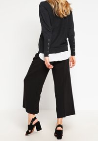 Maglione nero a maniche lunghe con dettagli a bottoni sulle maniche, indossato sopra una camicia bianca, abbinato a pantaloni wide-leg neri e sandali con tacco.