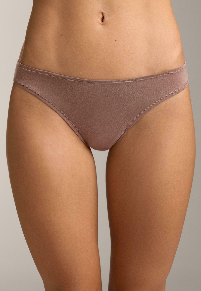 Frau trägt mauvefarbenes Baumwoll-Bikini-Unterwäsche, gezeigt von der Taille bis zur Mitte des Oberschenkels vor neutralem Hintergrund.