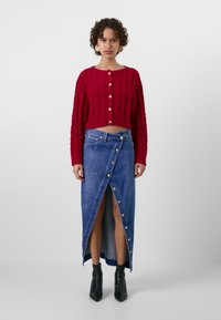 Pull en tricot rouge court avec un col rond et une fermeture à boutons, associé à une jupe portefeuille en denim bleu avec des boutons dorés et une fente. Bottines noires.