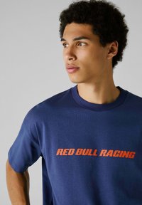 Tamsiai mėlynas medvilninis marškinėliai trumpomis rankovėmis ir apvalia kaklo iškirpte, su oranžiniu „RED BULL RACING“ grafiku priekyje.