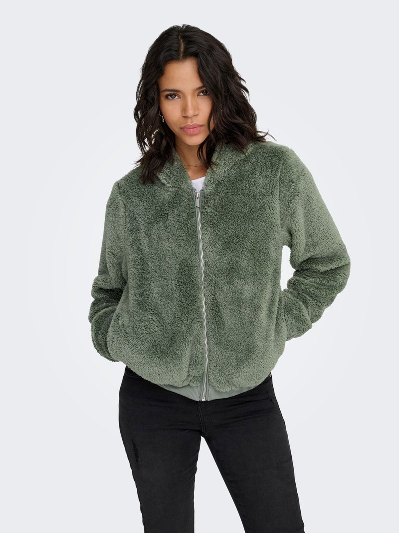 Veste zippée en velours vert avec une texture douce, des poignets et un ourlet côtelés. Style avec des poches latérales, associée à un pantalon noir ajusté.