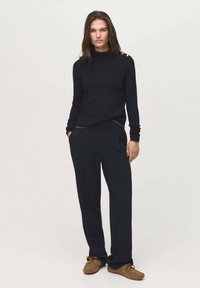 Black knitted sweater with a high neck and three accent buttons on the shoulder; paired with loose-fitting black trousers and brown shoes.