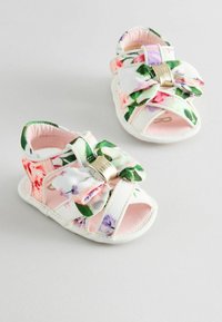 Kukka-aiheiset vauvan sandaalit valkoisella pohjalla, joissa on suuri, pehmeä nauha ja vaaleanpunainen vuori. Remmit on koristeltu vihreillä lehtikuoseilla ja kultaisilla yksityiskohdilla.
