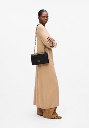 Sac à main en cuir noir avec une chaîne dorée, arborant un logo et un design structuré. Porté avec une longue robe beige et des ballerines à imprimé animal.