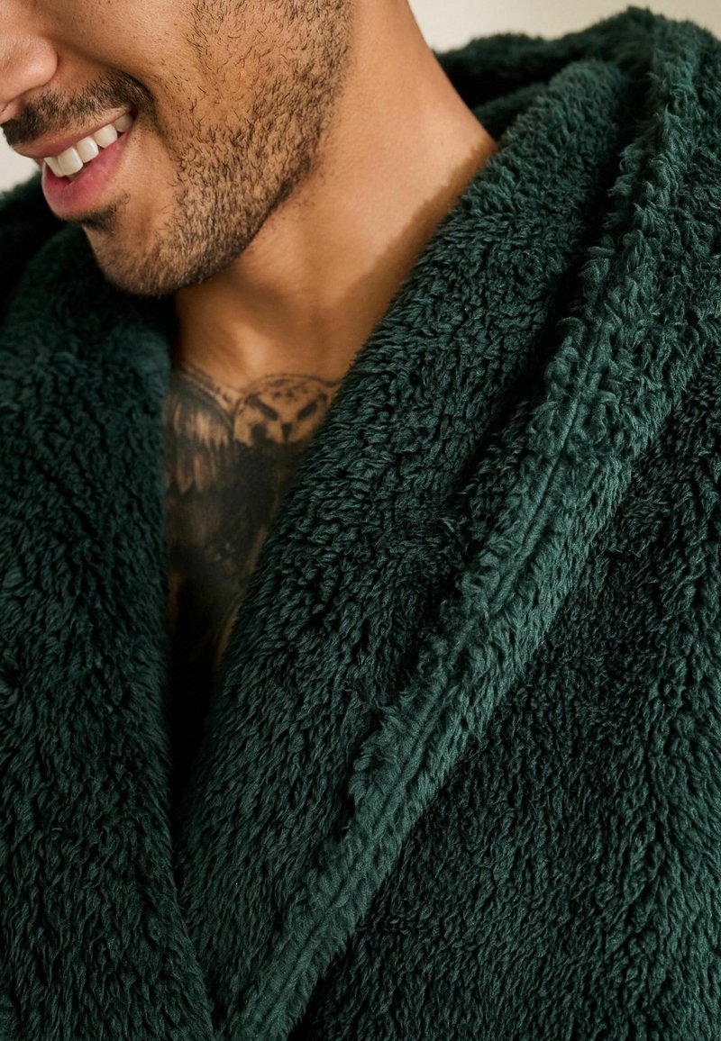 Uomo con barba che sorride, indossando un accappatoio di velluto verde scuro, che mostra un tatuaggio sul petto di un gufo con le ali spiegate.