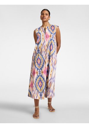 Mujer de pie con las manos detrás de la espalda, vistiendo un vestido sin mangas, hasta el tobillo, con patrones geométricos abstractos y coloridos y sandalias.