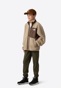 Beežide ja pruunide fliisjope lukuga, ees taskutega, kombineeritud oliivroheliste jooksupükste ja mustade matkasaabastega. Müts on pruuni korduroo materjalist.