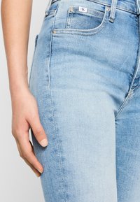 Šviesiai mėlyni džinsai su maža balta Calvin Klein logotipo etikete šalia priekinių kišenių ir ranka, ilsinčia šalia šlaunies.