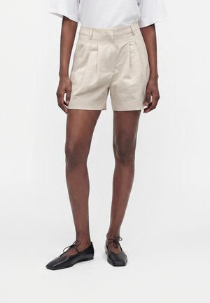 Person iført beige højtaljede plisserede shorts, hvid top og sorte balletsko med firkantet tå og ankelbind, stående mod en ensfarvet baggrund.