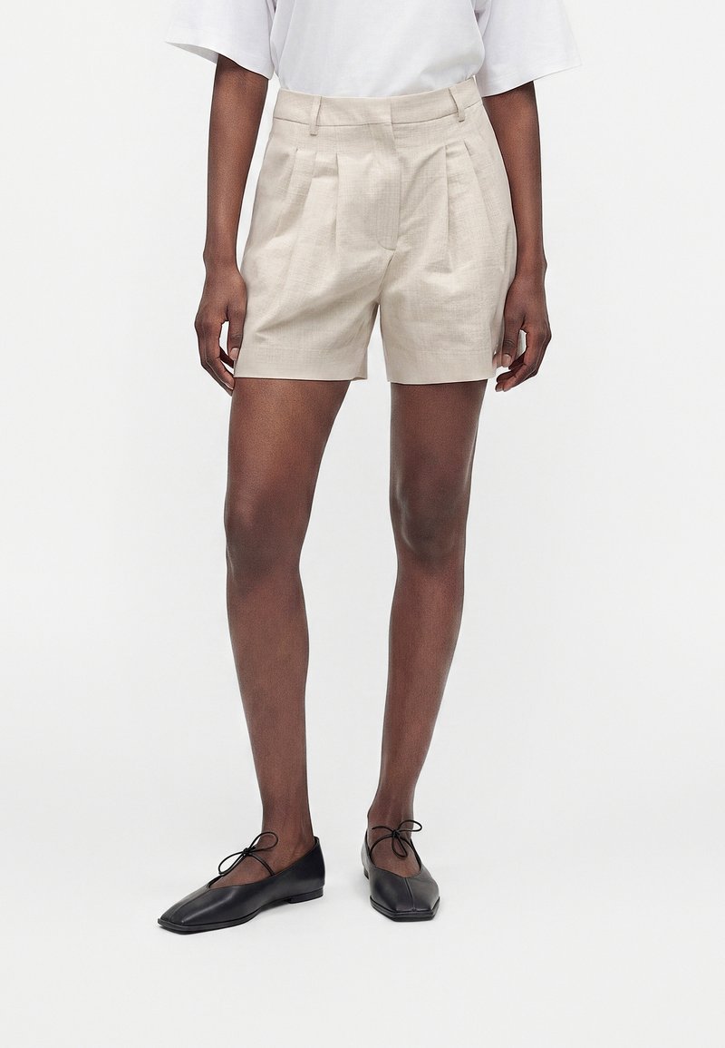 Personne portant un short plissé beige taille haute, un haut blanc et des ballerines noires à bout carré avec liens à la cheville, debout devant un fond uni.