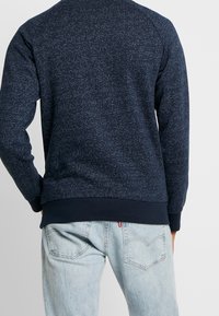 Hombre con sudadera azul marino moteada y jeans azul claro, visto desde atrás con las manos en los bolsillos, contra un fondo liso.