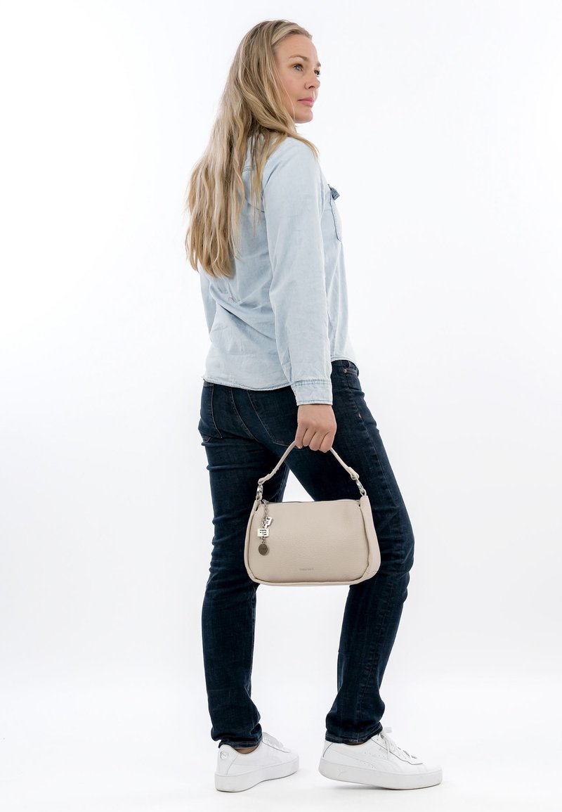 Vrouw met lang blond haar, draagt een lichtblauw shirt, donkere spijkerbroek, witte sneakers, houdt een beige handtas met metalen bedel vast, staand opzij.