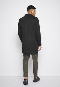 Abrigo negro con un diseño entallado y solapa de pico, combinado con pantalones marrones con estampado y zapatillas negras. Textura suave y forma ajustada.